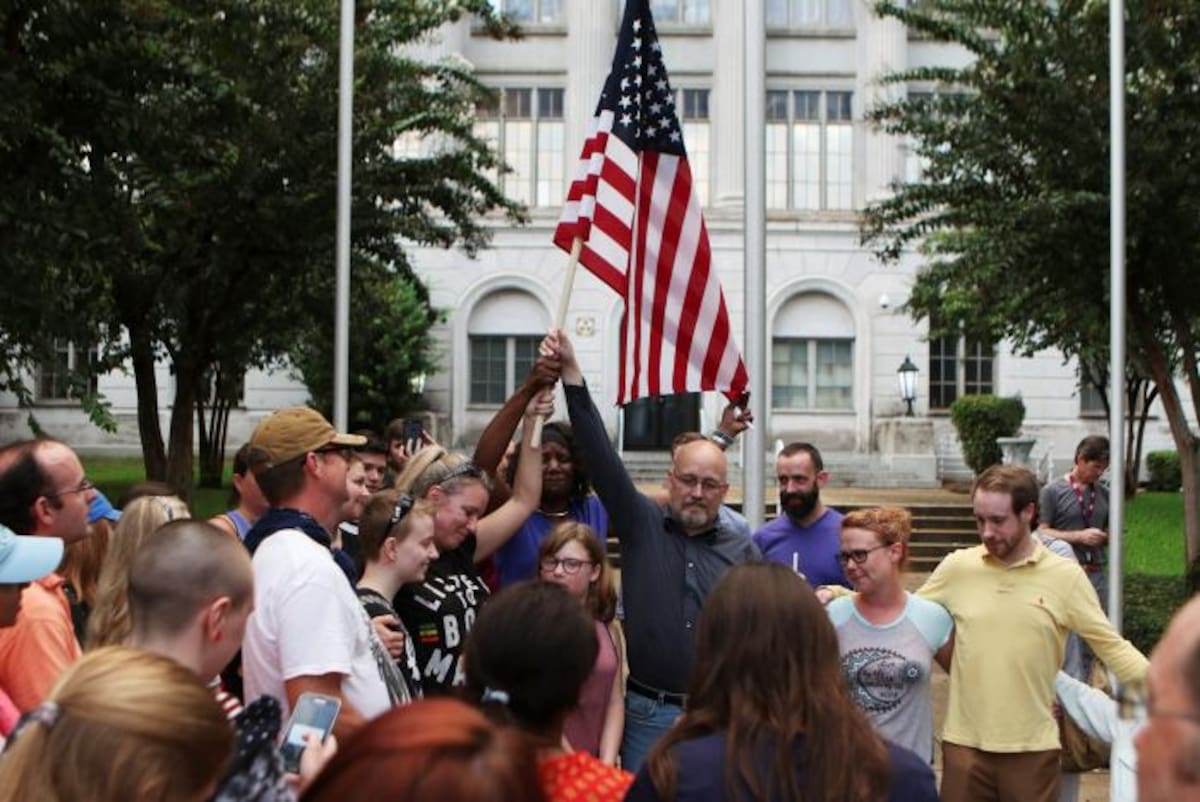 Los miembros de la comunidad que asisten a una vigilia rodean a Melissa Jeanne Byer, Marie Brown y Greg Sherburne cuando levantan una bandera estadounidense sobre el exterior del Palacio Parroquial de Ouachita en Monroe.